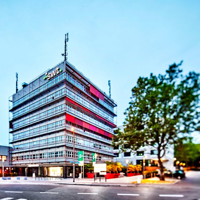 Ein moderner Bürokomplex mit einer Glasfassade in verschiedenen Farbtönen, darunter rot und grau. Das Gebäude hat mehrere Stockwerke und ist umgeben von Bäumen und Strassenlaternen. Im Vordergrund gibt es eine gepflasterte Fläche, auf der sich Fahrradständer befinden. Im Hintergrund sind weitere moderne Gebäude sichtbar, und die Umgebung scheint gut beleuchtet zu sein. Der Himmel ist klar und zeigt eine Dämmerungsstimmung.