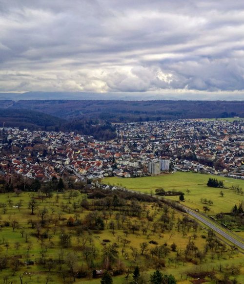 Eine Luftaufnahme von Dettenhausen im Landkreis Tübingen mit Wohnhäusern und Gewerbegebiet, umgeben von Feldern und Wäldern. Wolkenbedeckter Himmel.