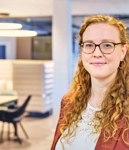 Eine Frau mit lockigem, langehaarigem Haar trägt eine Brille und ein braunes Jacket. Im Hintergrund ist ein Büro sichtbar.