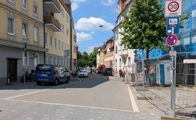 Eine Straßenszene mit Wohngebäuden, fahrenden Autos, Fahrrädern, Fußgängern und Baugerüst an der Seite