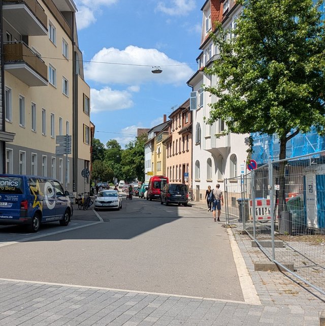 Eine Straßenszene mit Wohngebäuden, fahrenden Autos, Fahrrädern, Fußgängern und Baugerüst an der Seite