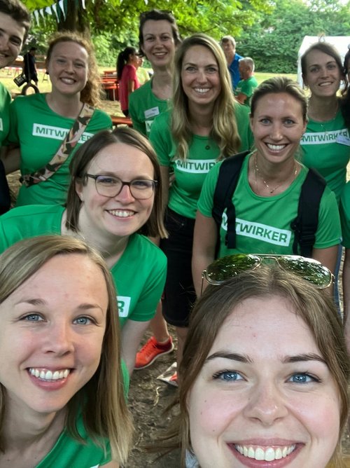 Eine Gruppe von Menschen in grünen T-Shirts mit dem Aufdruck "MITWIRKER" posiert fröhlich für ein Gruppenfoto im Freien. Die gesamte Szene wirkt entspannt und freundlich, umgeben von Bäumen und einer Sommeratmosphäre.