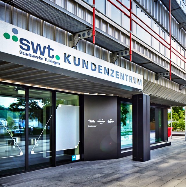 Ein modernes Gebäude mit einem Eingang, der mit dem Schriftzug „swt. Stadtwerke Tübingen KUNDENZENTRUM“ beschriftet ist. Die Fassade besteht aus Metall und hat große Fenster, die teilweise mit dekorativen Elementen versehen sind. Im Vordergrund sind Pflastersteine zu sehen und auf der linken Seite stehen einige Fahrräder an einem Abstellplatz. Links im Hintergrund sind Bäume und weitere Gebäude sichtbar. Die Szene ist hell beleuchtet und es ist ein sonniger Tag.
