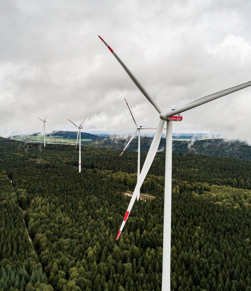 Eine Luftaufnahme von mehreren Windkraftanlagen, die zwischen dichten, grünen Wäldern stehen. Die Windräder sind von oben zu sehen, und ihre Rotorblätter sind in Bewegung. Der Himmel ist bewölkt, mit einigen Lichtreflexionen auf den Klingen. Im Hintergrund erheben sich sanfte Hügel, die von bewaldeten Flächen und Wiesen durchzogen sind.