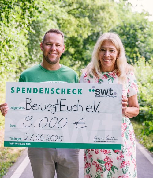 Zwei Personen stehen auf einem Weg umgeben von Pflanzen und Bäumen, sie halten einen großen Scheck über 9.000 Euro.