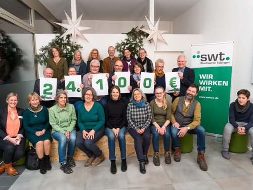 Eine Gruppe von Menschen sitzt und steht in einem Raum, umgeben von dekorativen Weihnachtssternen und Tannen. Sie halten große weiße Plakate mit grünen Zahlen, die zusammen 24.100 € ergeben. Auf der rechten Seite steht ein Banner mit der Aufschrift "swt. Stadtwerke Tübingen. Wir wirken mit." Die Anwesenden tragen verschiedene legere Kleidung und lächeln freundlich in die Kamera.