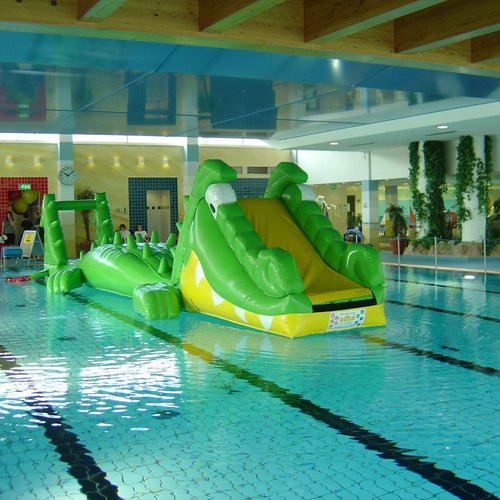 Ein großer, grüner, aufblasbarer Krokodilrutsche steht im Wasser eines Hallenbads neben bunten Luftballons und Stühlen
