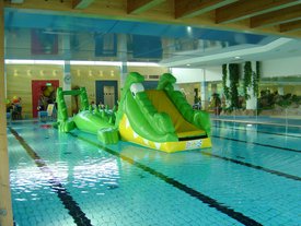 Ein großer, grüner, aufblasbarer Krokodilrutsche steht im Wasser eines Hallenbads neben bunten Luftballons und Stühlen