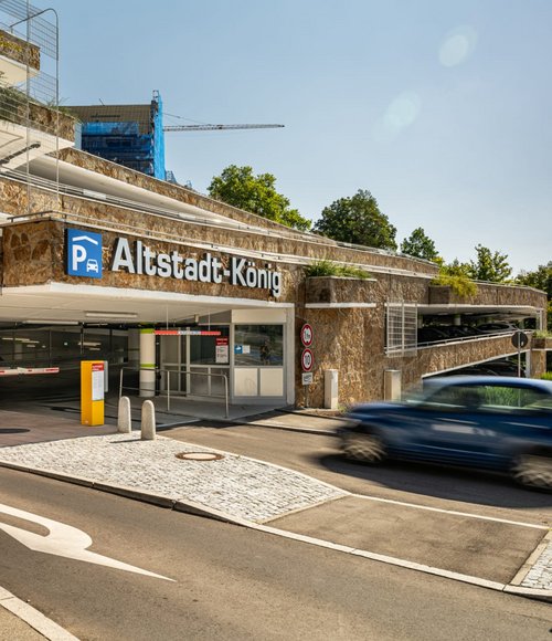 Ein blaues Auto fährt in das moderne Parkhaus „Altstadt-König“ in Tübingen ein. Die Fassade besteht aus Naturstein, das Gebäude ist begrünt, und eine Schrankenanlage regelt die Zufahrt.