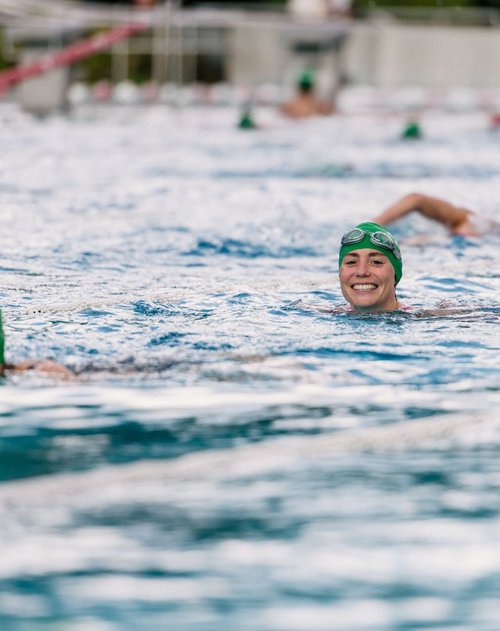 Eine Gruppe von Schwimmern in einem Schwimmbecken. Im Vordergrund schwimmt eine fröhlich lächelnde Sportlerin mit einer grünen Schwimmkappe und Schwimmbrille. Im Hintergrund sind weitere Schwimmer zu sehen, die ebenfalls grüne Kappen tragen. Das Wasser des Beckens spiegelt den Himmel und erzeugt sanfte Wellen.