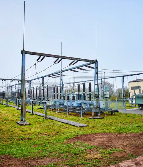 Eine Freiluft-Transformatorenstation mit mehreren Hochspannungsleitungen und Transformatoren. Im Hintergrund ist ein Gebäude mit Fenstern zu sehen, während die Umgebung von einer grünen Wiese umgeben ist. Der Himmel ist bewölkt und es herrscht eine ruhige Atmosphäre.