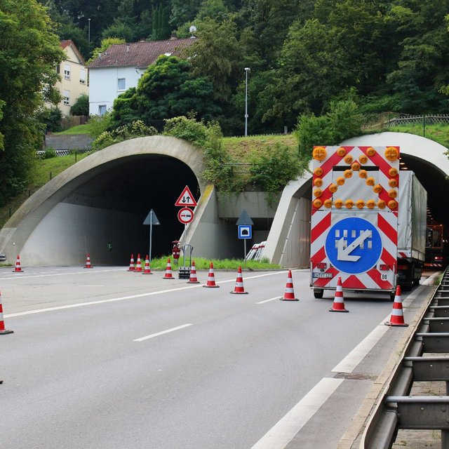 Eine Straße führt zu einem Tunnel mit zwei Einfahrten. Vor dem Tunnel stehen mehrere Verkehrshütchen und ein gelbes Fahrzeug mit Leuchtsignalen. Ein Verkehrszeichen zeigt eine Geschwindigkeitsbegrenzung von 30 km/h an. Im Hintergrund sind Bäume und einige Häuser sichtbar. An der Seite der Straße verläuft eine Schutzplanke.