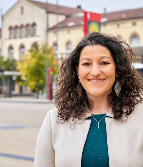 Eine fröhliche Frau mit lockigem, dunklem Haar steht in einer städtischen Umgebung. Sie trägt eine graue Jacke über einem grünen Oberteil und hat große, silberne Ohrringe. Im Hintergrund sind Gebäude, eine Straßenbahn und einige Passanten zu sehen. Es ist bewölkt und die Szene wirkt lebhaft.