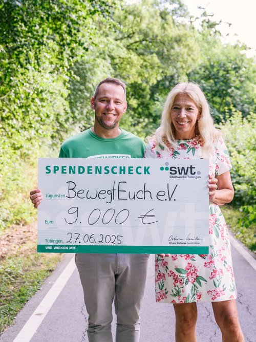 „SPENDENSCHECK zugunsten BewegEuch e.V. 9.000 €“, sowie das Datum „27.06.2025“ und der Hinweis „Stadtwerke Tübingen“. Die Person links trägt ein grünes T-Shirt, während die Person rechts ein Kleid mit Blumenmuster trägt.