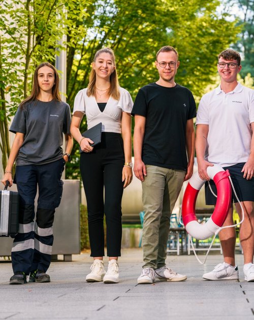 eine Person hält einen Koffer, eine andere hat ein Tablet in der Hand, während ein weiterer Teilnehmer einen Rettungsring und eine Bohrmaschine hält. Die Kleidung reicht von lässig bis sportlich, und die Gruppenmitglieder scheinen eine freundliche und entspannte Atmosphäre auszustrahlen.