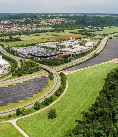 Eine Luftaufnahme zeigt ein Gebiet mit Solarpanelen und einer angrenzenden Industrie- und Gewerbezone. Auf der linken Seite sind einige Firmengebäude und Parkplätze sichtbar, während sich rechts ein weitläufiges Feld erstreckt. In der Mitte verlaufen Straßen und Kurven, die durch die grünen Wiesen und Wälder ergänzt werden. Im Hintergrund sind Wohngebäude und weitere Landschaftselemente zu sehen, die Teil einer städtischen Umgebung sind.