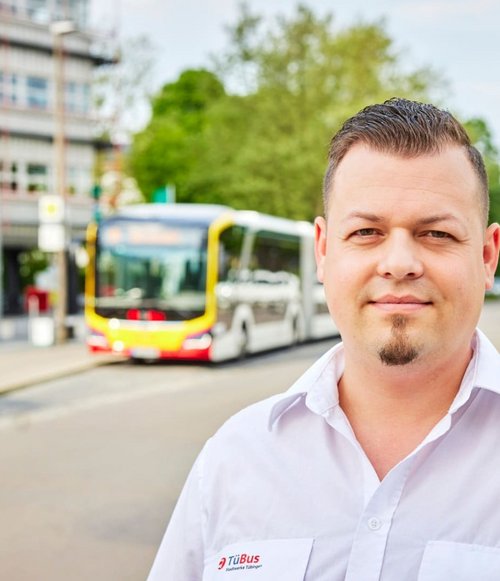 Ein Mann mit kurzen, dunklen Haaren und einem weißen Hemd steht auf einer Straße. Im Hintergrund sind zwei Busse und Bäume sichtbar.