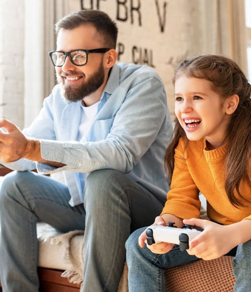 Ein Mann und ein Kind sitzen auf einem Sofa und spielen Videospiele. Der Mann trägt eine Brille und hat einen Bart. Das Kind, ein Mädchen mit langen Haaren, trägt einen orangefarbenen Pullover und lächelt. Beide halten Videospiel-Controller in den Händen und schauen konzentriert auf den Bildschirm. Im Hintergrund sind Fenster mit Tageslicht und eine Wand mit einzigartigen Markierungen zu sehen.
