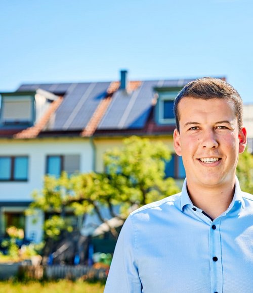 Ein junger Mann mit kurzem Haar trägt ein hellblaues Hemd, im Hintergrund eine Wohnsiedlung mit Solaranlagen und Bäumen.