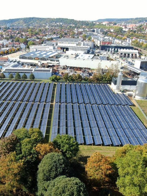 Eine Luftaufnahme einer großen Solarpark-Anlage, die aus mehreren Reihen von Solarpanelen besteht. Die Solarpanels sind in gleichmäßigen, schrägen Reihen angeordnet und bedecken eine grüne Wiese, die von Bäumen umgeben ist. Im Hintergrund sieht man eine städtische Umgebung mit Wohngebäuden, Geschäften und Straßen. Die Landschaft ist von herbstlichen Farben geprägt, mit bunten Blättern an den Bäumen. In der Nähe der Solaranlage befindet sich ein silberner Speicherbehälter und einige industrielle Gebäude.