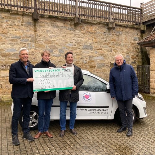 Vier Personen stehen neben einem weißen Auto, das einen Aufkleber der Sozialen Dienste trägt. Sie halten einen großen Scheck mit dem Betrag von 3.900 Euro hoch. Im Hintergrund ist eine Steinmauer zu sehen. Die Personen schauen freundlich in die Kamera. Es scheint, als sei die Szene im Freien aufgenommen worden, möglicherweise an einem verregneten oder bewölkten Tag.