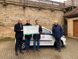 Vier Personen stehen neben einem weißen Auto, das einen Aufkleber der Sozialen Dienste trägt. Sie halten einen großen Scheck mit dem Betrag von 3.900 Euro hoch. Im Hintergrund ist eine Steinmauer zu sehen. Die Personen schauen freundlich in die Kamera. Es scheint, als sei die Szene im Freien aufgenommen worden, möglicherweise an einem verregneten oder bewölkten Tag.
