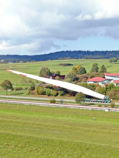 Eine große Windkraftanlagenblatt wird auf einem Lastwagen transportiert, während im Hintergrund eine ländliche Landschaft mit grünen Feldern, Bäumen und Hügeln sichtbar ist. Einige Häuser mit roten Dächern liegen am Straßenrand, und eine zweitekante auf dem Weg ist ebenfalls zu sehen. Der Himmel ist teilweise bewölkt.