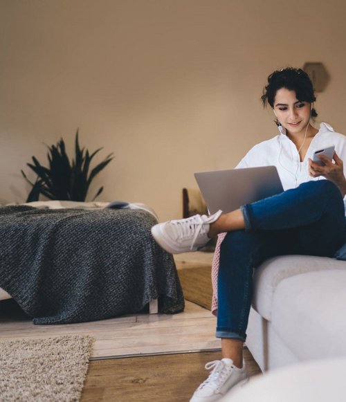 Frau sitzt auf einem Sofa mit Laptop und Smartphone, lächelnd bei der Nutzung beider Geräte.