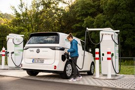 Eine Person tankt ein weißes Elektroauto, das ein VW ID. Buzz ist, an einer Ladestation. Die Ladestation steht auf einem gepflasterten Bereich und befindet sich in einer bewaldeten Umgebung. Im Hintergrund sind Bäume und eine grüne Landschaft sichtbar. Die Person trägt eine blaue Jacke und hat braune Haare, die nach hinten gebunden sind.