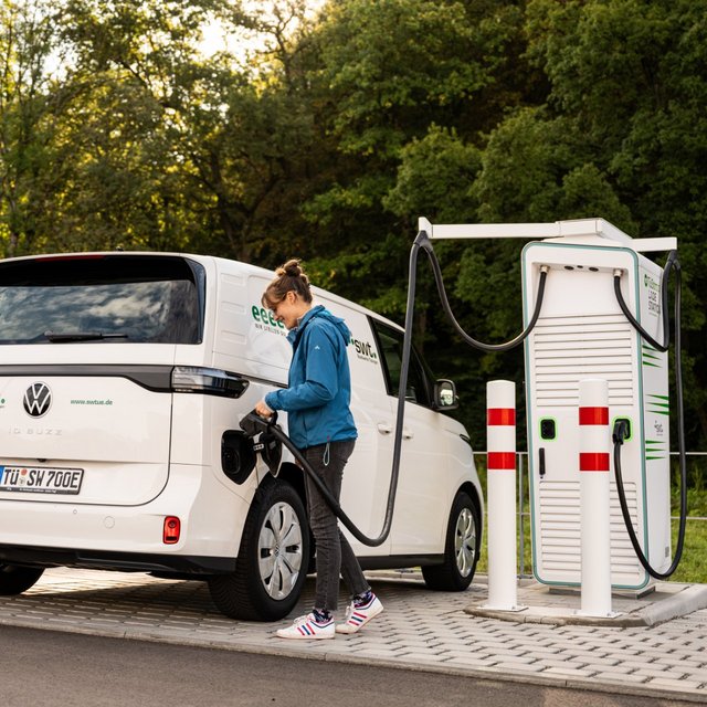 Eine Person tankt ein weißes Elektroauto, das ein VW ID. Buzz ist, an einer Ladestation. Die Ladestation steht auf einem gepflasterten Bereich und befindet sich in einer bewaldeten Umgebung. Im Hintergrund sind Bäume und eine grüne Landschaft sichtbar. Die Person trägt eine blaue Jacke und hat braune Haare, die nach hinten gebunden sind.