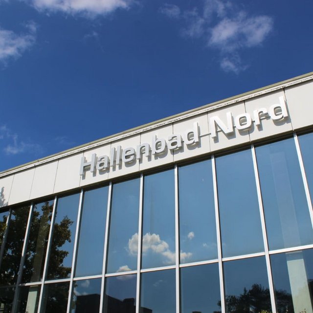 Ein modernes Gebäude mit großen Fenstern reflektiert den blauen Himmel und einige Wolken. Oben am Gebäude prangt der Schriftzug "Hallenbad Nord". Neben dem Gebäude sind einige Bäume zu sehen.