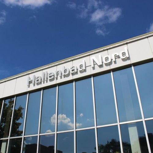Ein modernes Gebäude mit großen Fenstern reflektiert den blauen Himmel und einige Wolken. Oben am Gebäude prangt der Schriftzug "Hallenbad Nord". Neben dem Gebäude sind einige Bäume zu sehen.