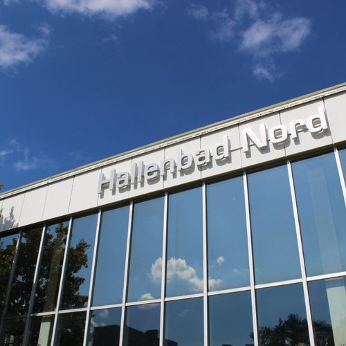 Ein modernes Gebäude mit großen Fenstern reflektiert den blauen Himmel und einige Wolken. Oben am Gebäude prangt der Schriftzug "Hallenbad Nord". Neben dem Gebäude sind einige Bäume zu sehen.