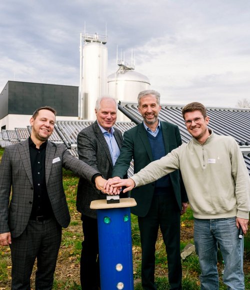 Vier Männer stehen mit ihren Händen auf einem blauen Knopf inmitten von Solarpanelen und großen Lagertanks