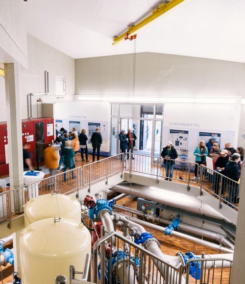 In einem großen Raum mit Maschinen stehen Besucher an Schaltern und informieren sich über die Ausstellung.