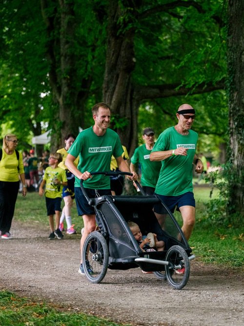 Zwei Männer in grünen T-Shirts joggen auf einem Weg durch einen Park, während einer von ihnen einen Kinderwagen schiebt. Im Hintergrund sind weitere Personen zu sehen, die ebenfalls laufen oder spazieren gehen. Die Umgebung ist von grünen Bäumen umgeben, und es herrscht eine lebhafte Atmosphäre.