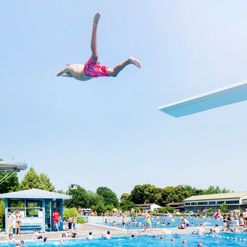Ein Junge springt im Badeanzug von einem Sprungbrett in ein Schwimmbecken voller Menschen und grüner Umgebung