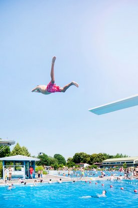 Ein Junge springt im Badeanzug von einem Sprungbrett in ein Schwimmbecken voller Menschen und grüner Umgebung