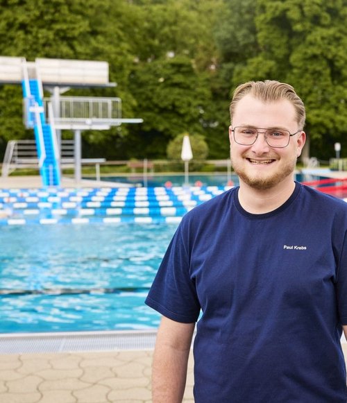 Ein junger Mann mit Brille und blauer T-Shirt steht am Rand eines Schwimmbades mit Wasser und Sprungbrett im Hintergrund