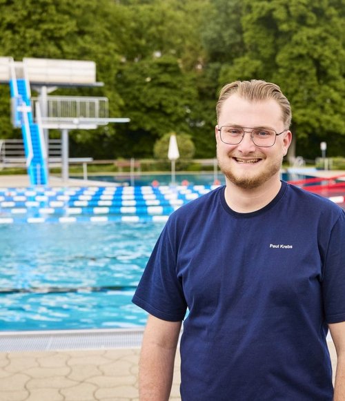 Ein junger Mann mit Brille und blauer T-Shirt steht am Rand eines Schwimmbades mit Wasser und Sprungbrett im Hintergrund