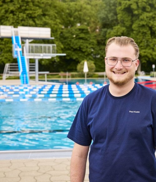 Ein junger Mann mit Brille und blauer T-Shirt steht am Rand eines Schwimmbades mit Wasser und Sprungbrett im Hintergrund