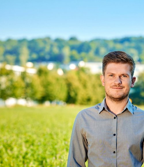 Ein Mann mit braunen Haaren trägt ein graues Hemd und steht in einem grünen Feld vor einer bewaldeten Landschaft.