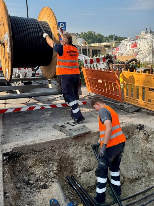 Zwei Arbeiter in orangefarbener Sicherheitskleidung stehen an einer Baustelle. Der eine ist dabei, ein großes Kabel auf einer Rolle von einem Fahrzeug abzunehmen, während der andere in einer Grube steht, die mit Kies gefüllt ist. Es sind auch mehrere Baustellenschilder und Barrieren sichtbar, die den Arbeitsbereich absichern. Im Hintergrund sind Baugeräte und eine aufgeschüttete Fläche mit Materialien zu sehen.