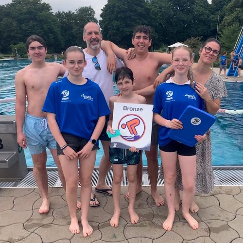 Sechs Personen stehen am Beckenrand eines Schwimmbades. Im Hintergrund sind ein Schwimmbecken und eine Rutsche sichtbar.