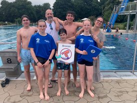 Sechs Personen stehen am Beckenrand eines Schwimmbades. Im Hintergrund sind ein Schwimmbecken und eine Rutsche sichtbar.