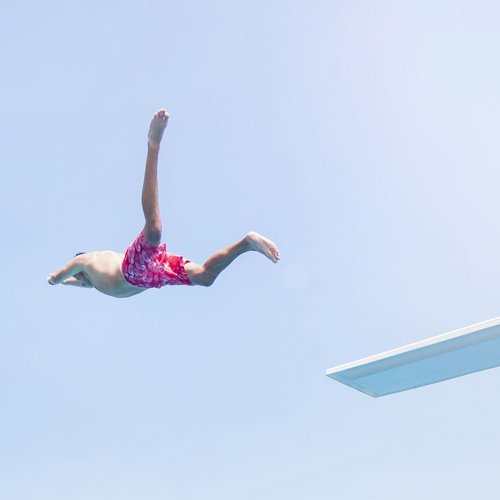 Ein Junge springt im Badeanzug von einem Sprungbrett in ein Schwimmbecken voller Menschen und grüner Umgebung