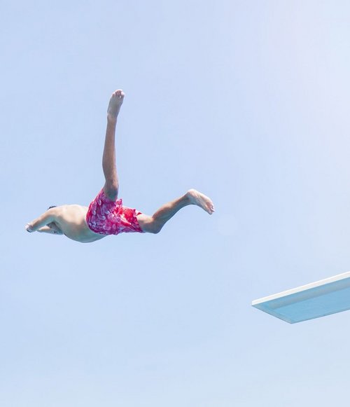 Ein Junge springt im Badeanzug von einem Sprungbrett in ein Schwimmbecken voller Menschen und grüner Umgebung