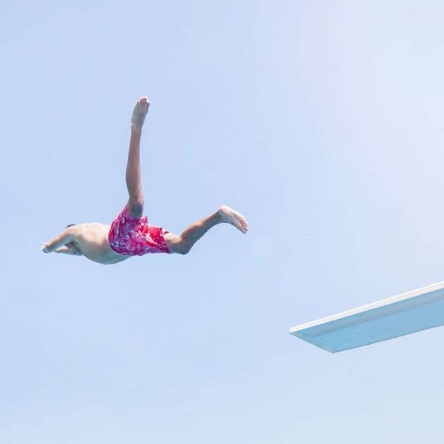 Ein Junge springt im Badeanzug von einem Sprungbrett in ein Schwimmbecken voller Menschen und grüner Umgebung