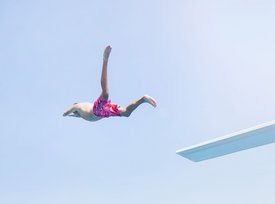Ein Junge springt im Badeanzug von einem Sprungbrett in ein Schwimmbecken voller Menschen und grüner Umgebung