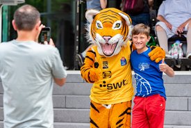 Ein Junge steht neben einem Tiger-Maskottchen, beide lächeln und posieren für ein Foto. Im Hintergrund sind Zuschauer sichtbar